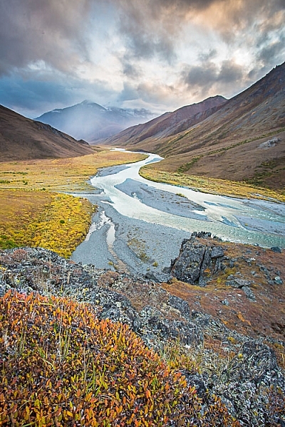 The autumn colors in the close foreground provide a good starting place for this image, guiding the eye to the winding river and then onto the stormy mountains beyond.